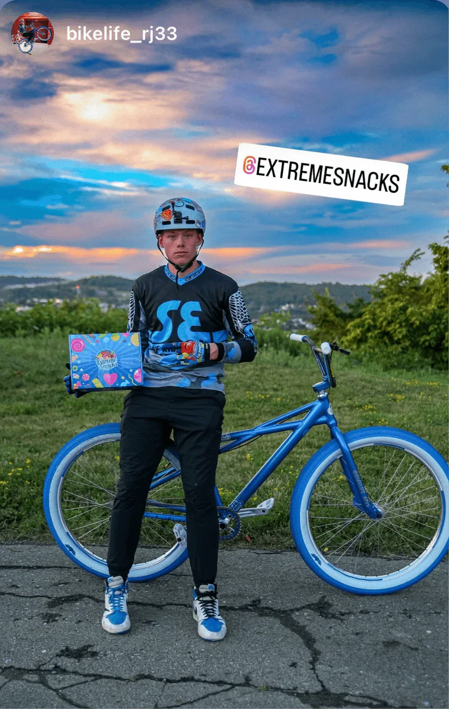 Person in cycling gear holding Extreme Snacks box with blue bike outdoors at sunset.