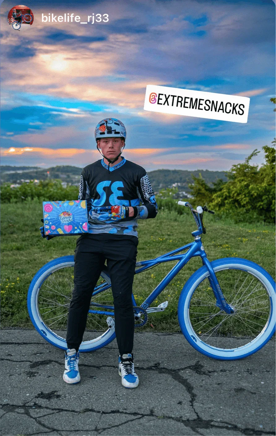 Man in cycling gear holding a box of exotic snacks next to a blue bike outdoors at sunset.