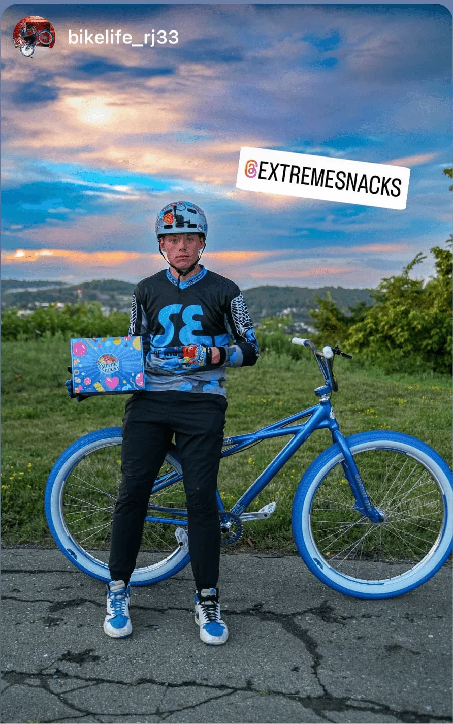 Biker in blue gear with a blue bike holding an Extreme Snacks box outdoors at sunset.